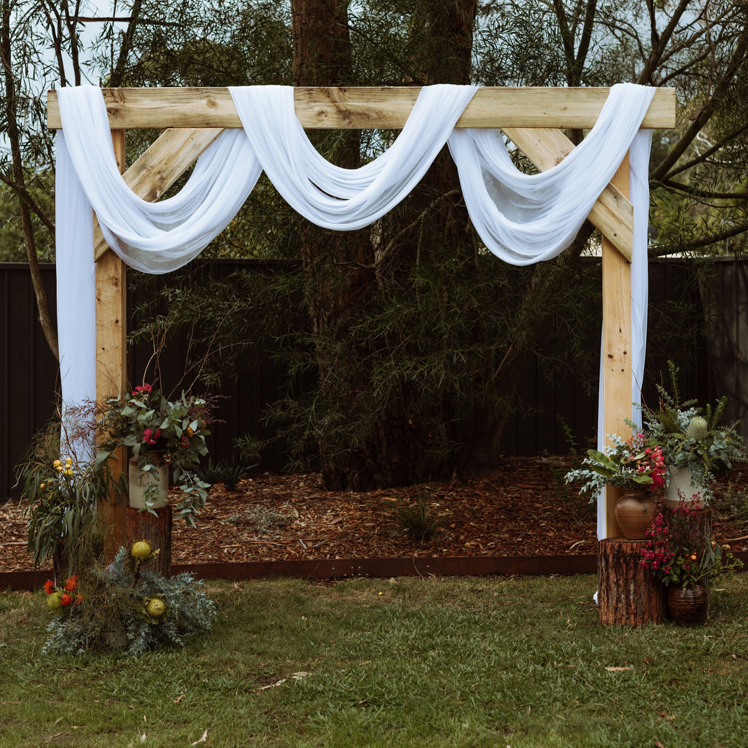 White sheer stretch chiffon draped over wooden arbor with native floral arrangements in front
