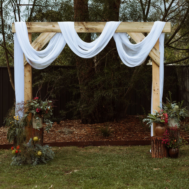 White sheer stretch chiffon draped over wooden arbor with native floral arrangements in front