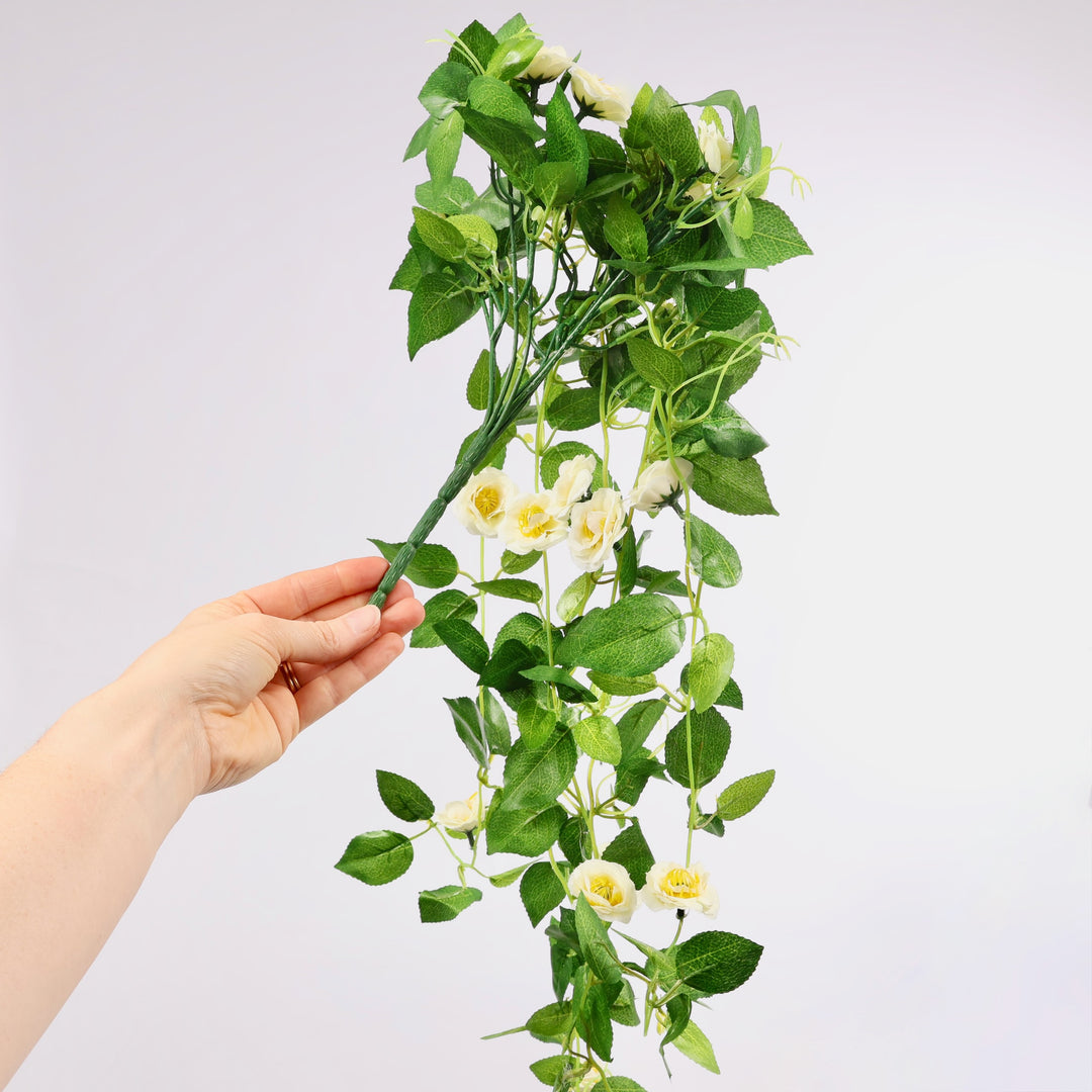 Hand holding a green artificial flower vine with white flowers against a light gray background