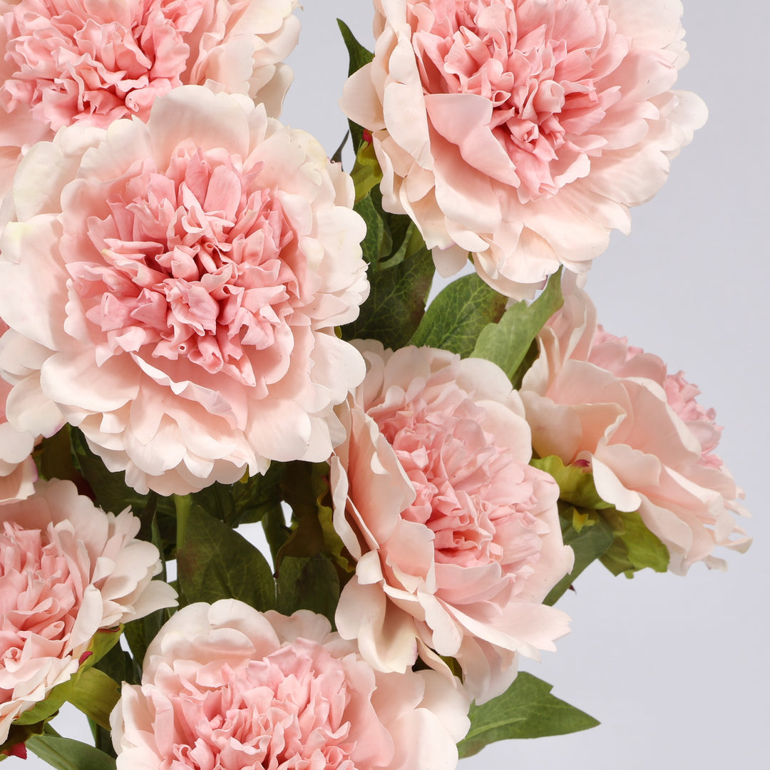 A close-up of a bouquet of artificial blush peony flowers with green leaves, set against a plain light gray background.