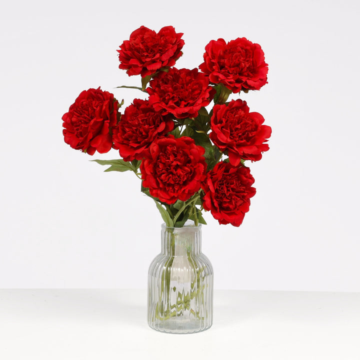 A bouquet of artificial red peony flowers with green stems in a clear ribbed glass vase, set against a plain white background.