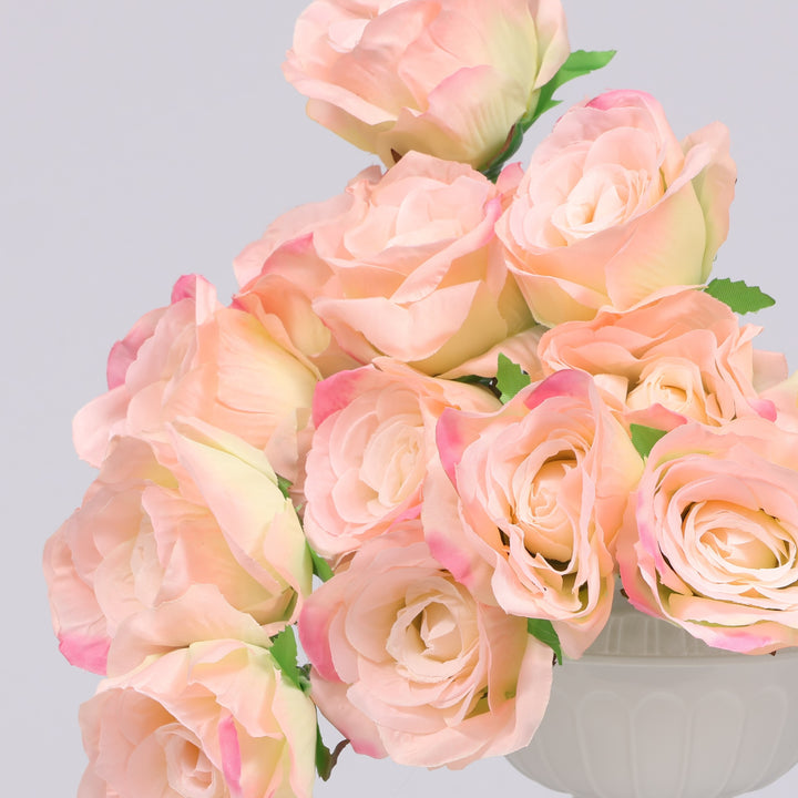 A bouquet of artificial pale pink roses with soft cream centers and green leaves, arranged in a white vase against a light gray background.