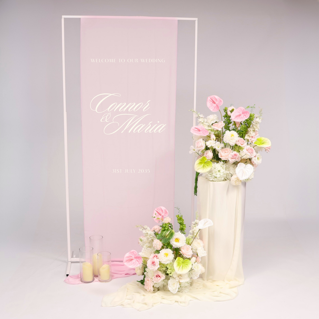 White Height Adjustable Sign, Light Pink Flower Arrangements with Anthurium Stems added, sitting on the White Round Plinth Set with Ivory Chiffon Table Runner, flickering LED pillar candles in Glass Vase Set of 3.