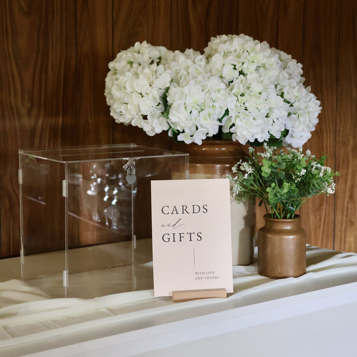 Clear wishing well sitting on a table with card and gift sign, hydrangeas, eucalyptus and baby's breath. 
