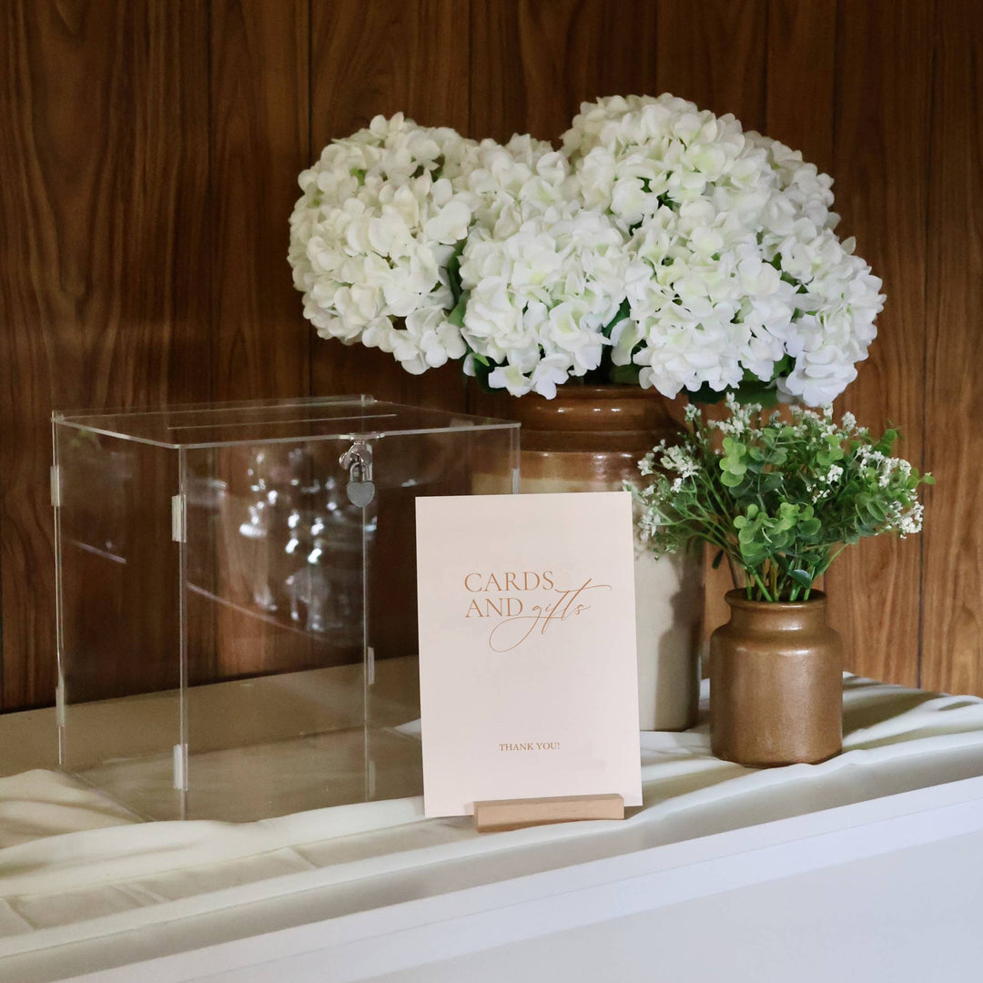 Card holder sitting on a table with sign for cards and gifts, in front of a clear wishing well with hydrangeas and eucalyptus arrangements