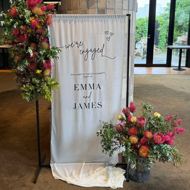 Black height adjustable frame with chiffon engagement sign and native Australian flowers around it. 