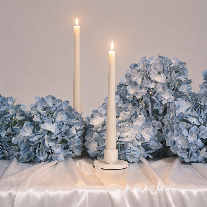 Two white candles in holders surrounded by blue hydrangeas on a white surface.