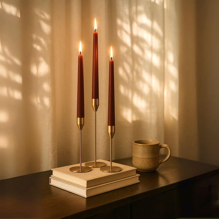 Ambient shot of taper candle holders sitting on top of a stack of books with lit  Burgundy taper candles. A mug sits to the right and there are sheer curtains behind with soft light filtered in