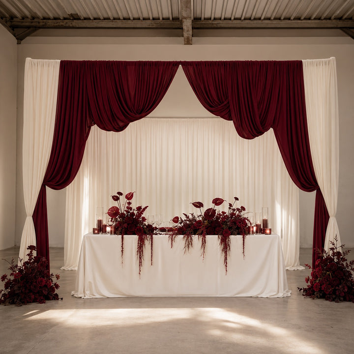 A formal event backdrop featuring deep burgundy draped curtains and white drapes, with a white draped table decorated with matching burgundy floral arrangements in a bright, modern hall.
