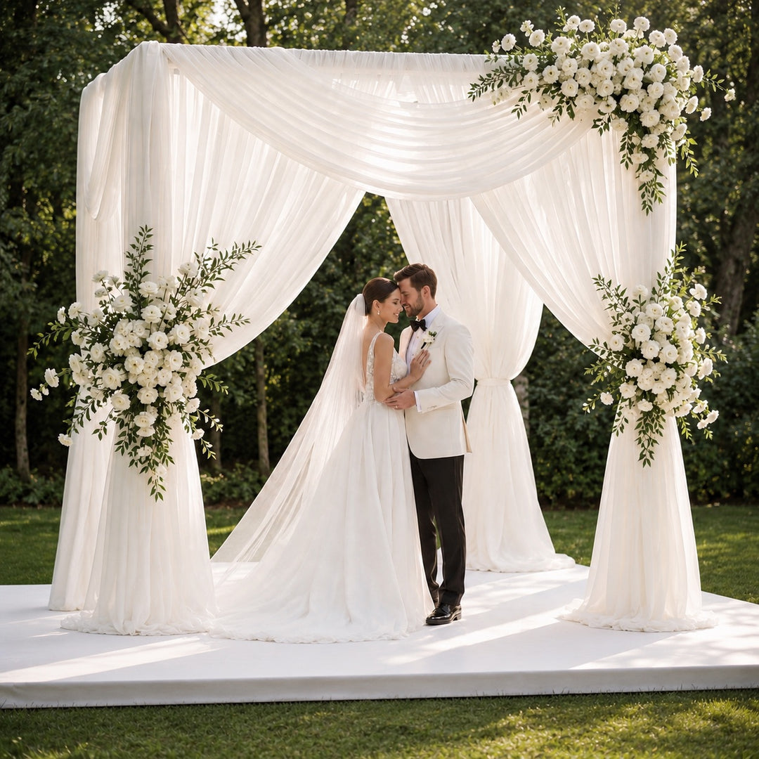 A bride and groom kiss under a white draped wedding canopy decorated with large white floral arrangements, set outdoors on a grassy lawn with trees in the background.