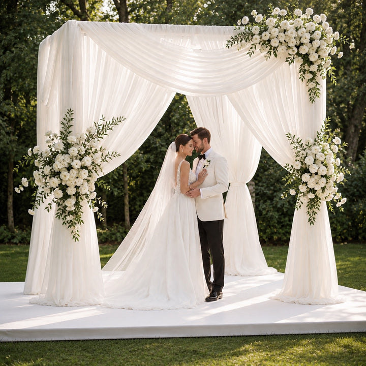 A bride and groom kiss under a white draped wedding canopy decorated with large white floral arrangements, set outdoors on a grassy lawn with trees in the background.