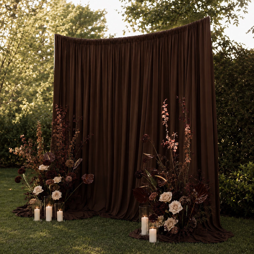 A large draped brown fabric backdrop set up outdoors on grass, with floral arrangements and lit candles placed in front, surrounded by trees and shrubs.