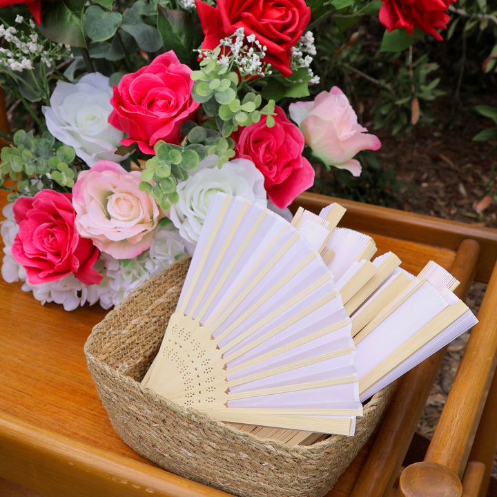 Paper hand fans in a woven basket, sitting next to flower arrangement of roses, peonies, eucalyptus and baby's breath