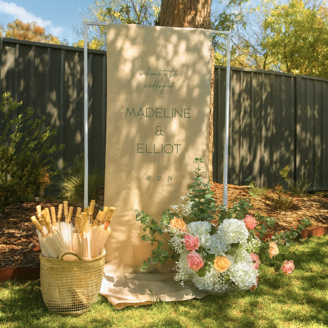 Height adjustable frame holding a linen welcome sign, with a flower arrangement and basket of paper parasols at the base. 