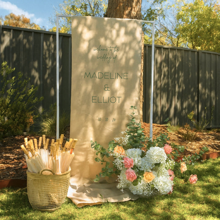 Height adjustable frame holding a linen welcome sign, with a flower arrangement and basket of paper parasols at the base. 