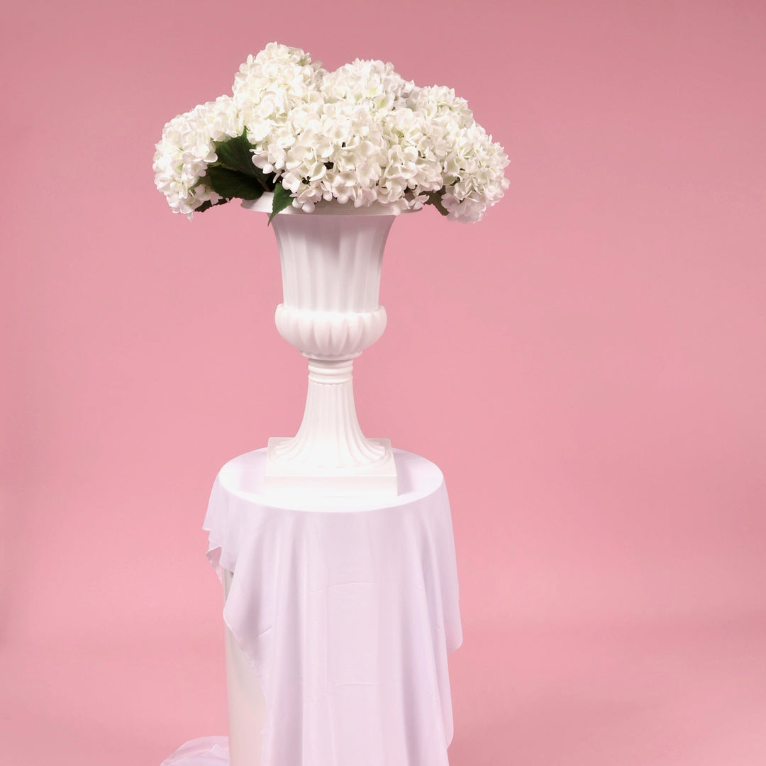 White hydrangea stems in a Trumpet vase, sitting on a white round plinth with a white chiffon runner in front of a pink backdrop