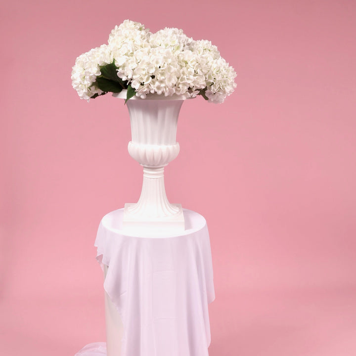 White hydrangea stems in a Trumpet vase, sitting on a white round plinth with a white chiffon runner in front of a pink backdrop