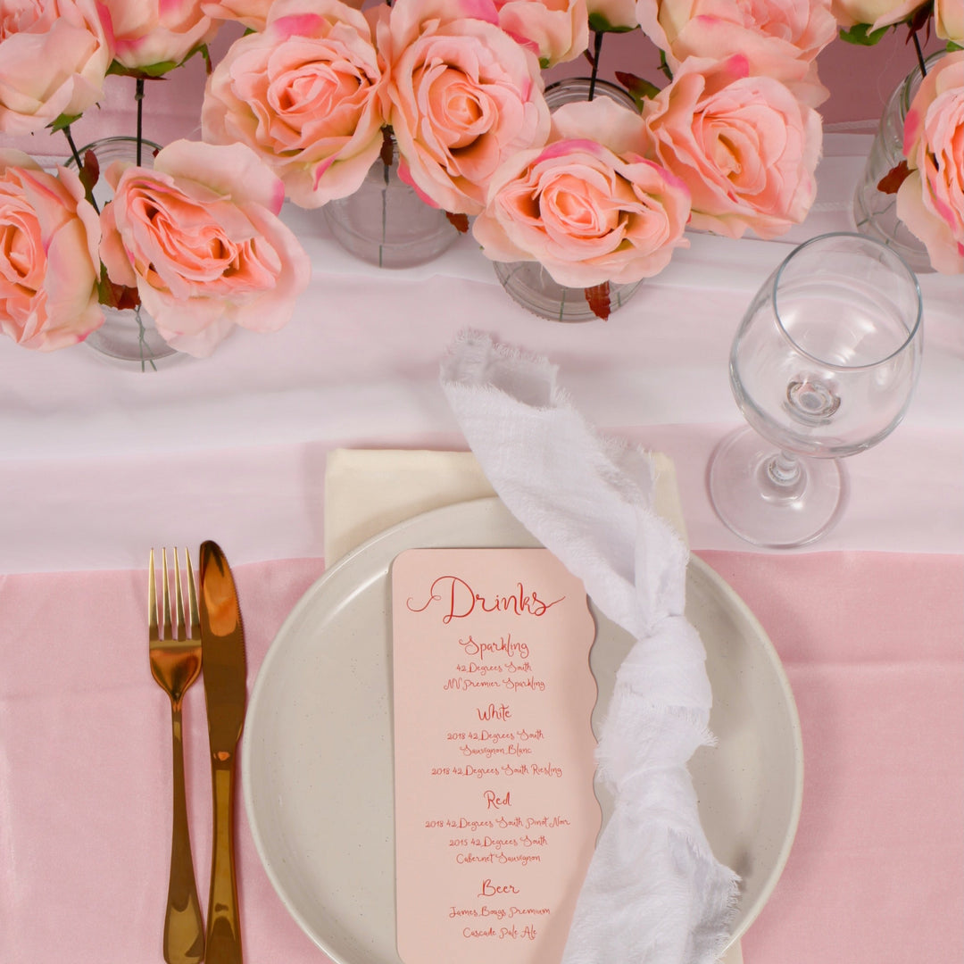 Blush peony flowers used as centerpiece on table with white chiffon runner and pink satin tablecloth. Gold cutlery and beige plate on table with a drinks menu sitting on top and a wine glass to the right.