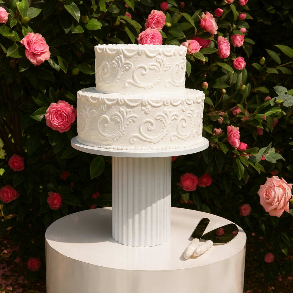 Decorated white cake on ribbed cake stand, on top of round plinth in front of garden with pink flowers.
