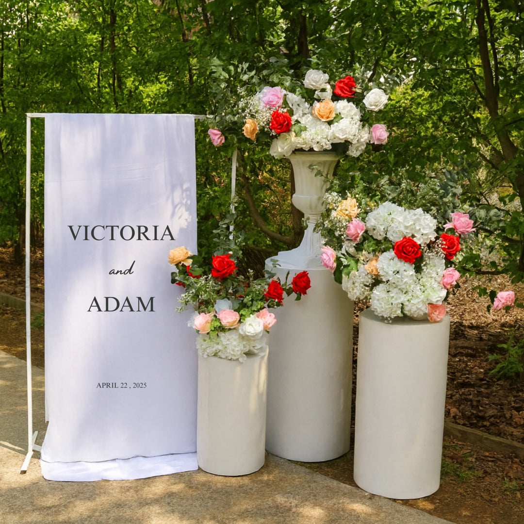 Welcome sign with white round plinth set and floral arrangements made from hydrangeas, roses (peach, blush, white, red and pink) eucalyptus and baby's breath, in front of garden