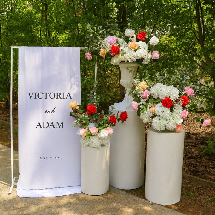 Welcome sign with white round plinth set and floral arrangements made from hydrangeas, roses (peach, blush, white, red and pink) eucalyptus and baby's breath, in front of garden