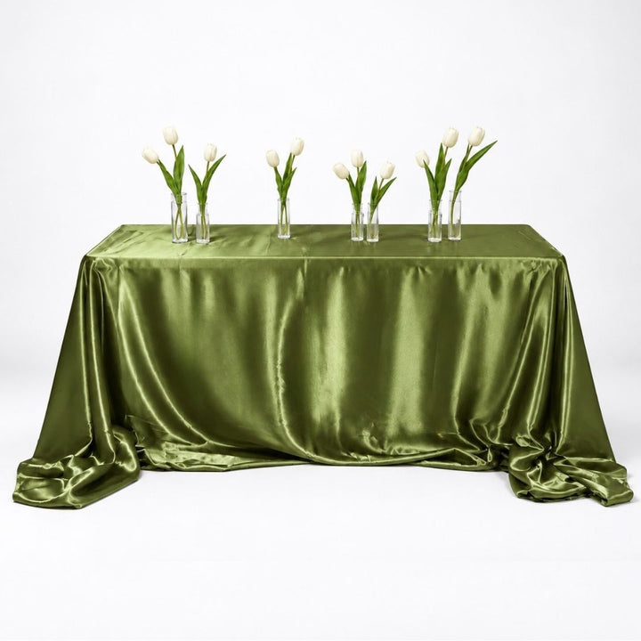 An olive green satin tablecloth draped over a rectangular table, styled with six clear glass vases containing white tulips, set against a seamless white studio background.