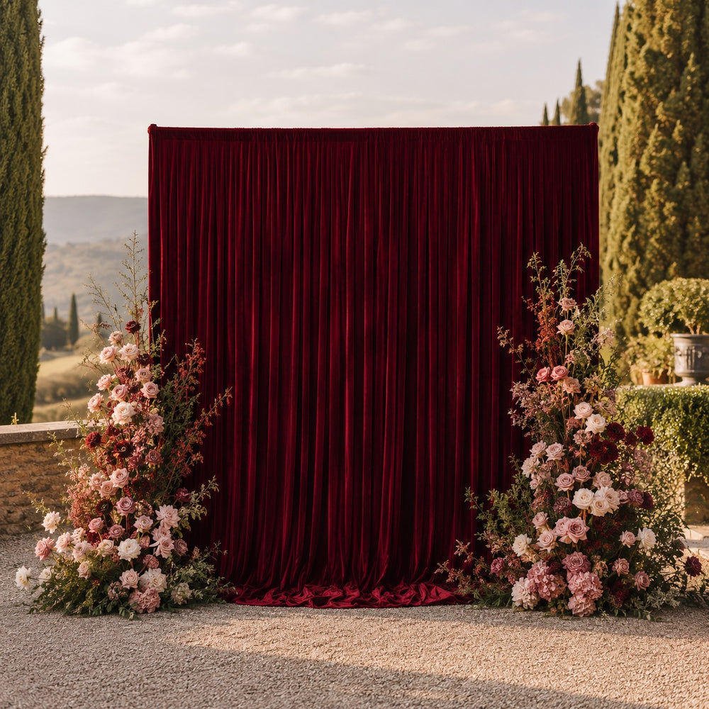 A burgundy velvet backdrop curtain set up outdoors, tall floral arrangements either side with pink, burgundy, and cream flowers, with a scenic landscape in the background.