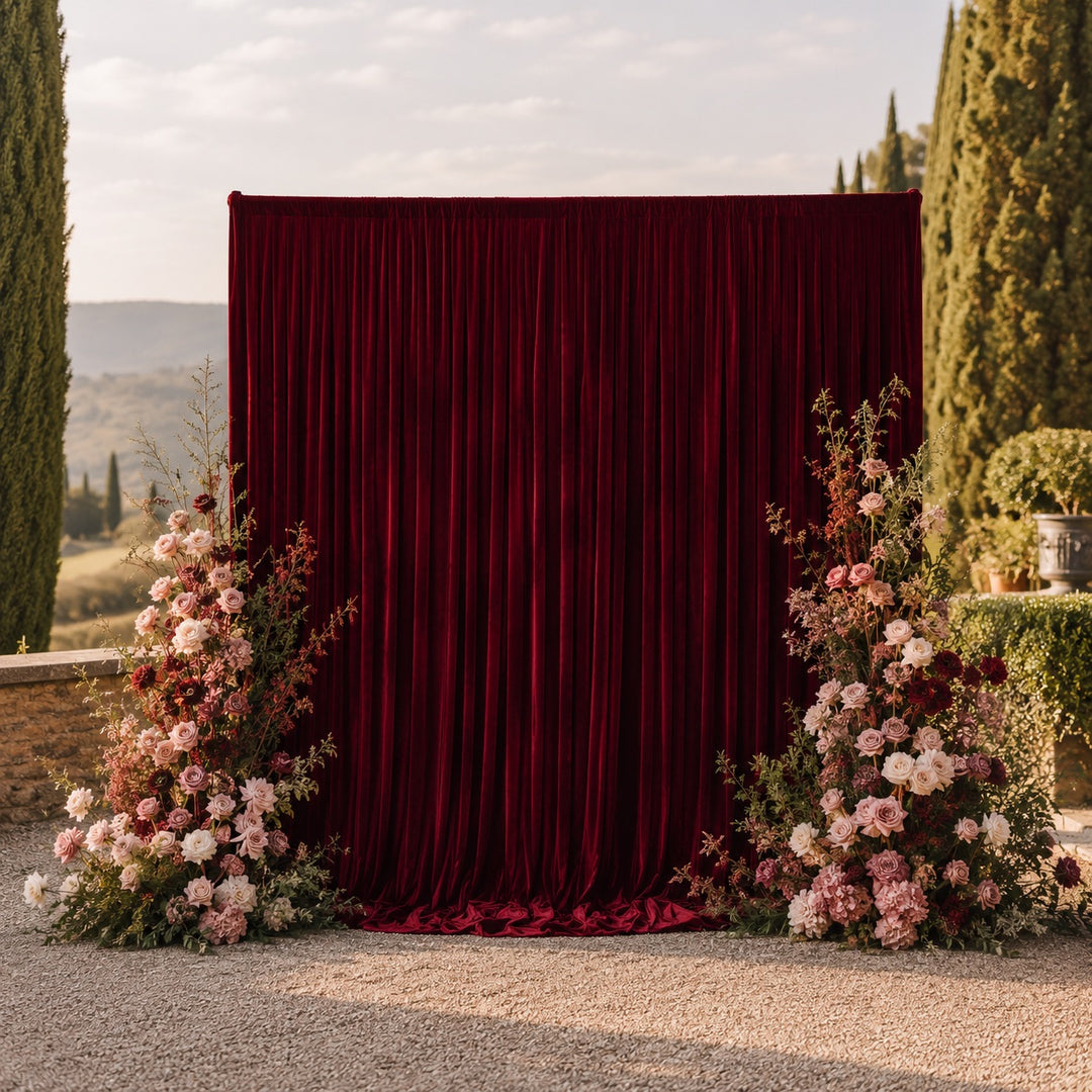 A burgundy velvet backdrop curtain set up outdoors, tall floral arrangements either side with pink, burgundy, and cream flowers, with a scenic landscape in the background.