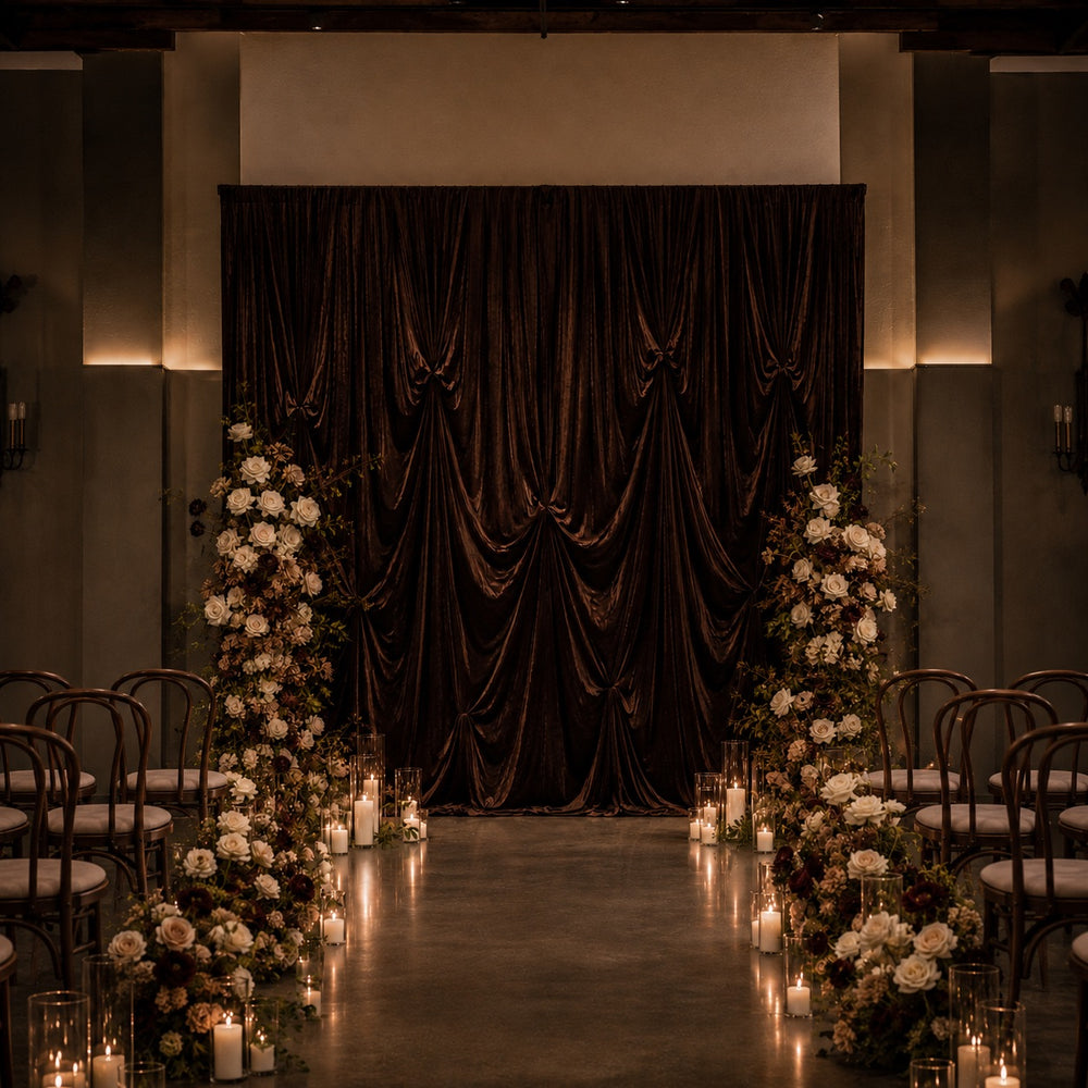 A dramatic event backdrop featuring a large draped dark brown velvet curtain, flanked by tall floral arrangements of white and brown flowers with lit candles on the floor, set against a dimly lit venue with rows of chairs.